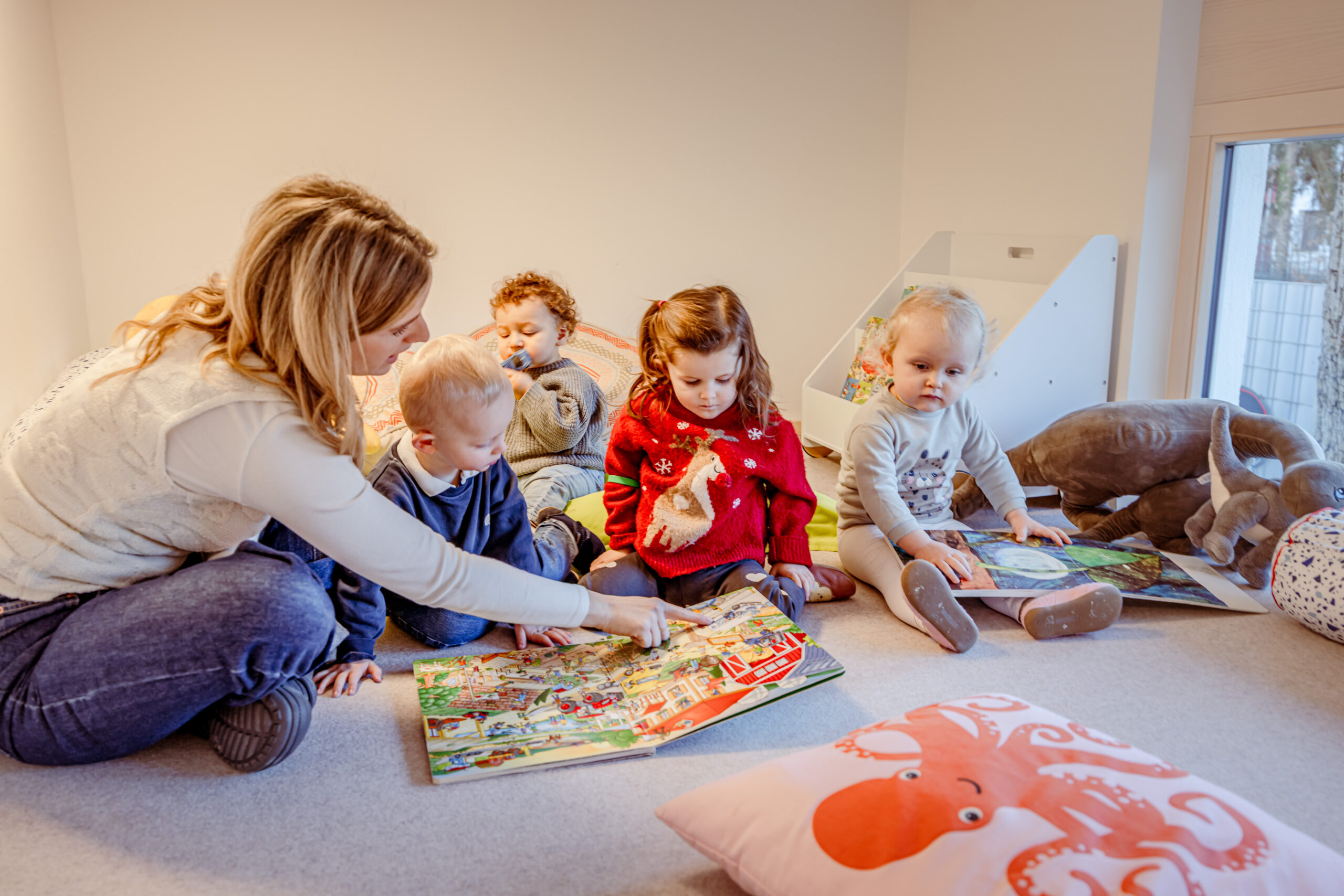 Eine Erzieherin spielt mit Kindern auf dem Boden und liest mit ihnen gemeinsam ein Buch.
